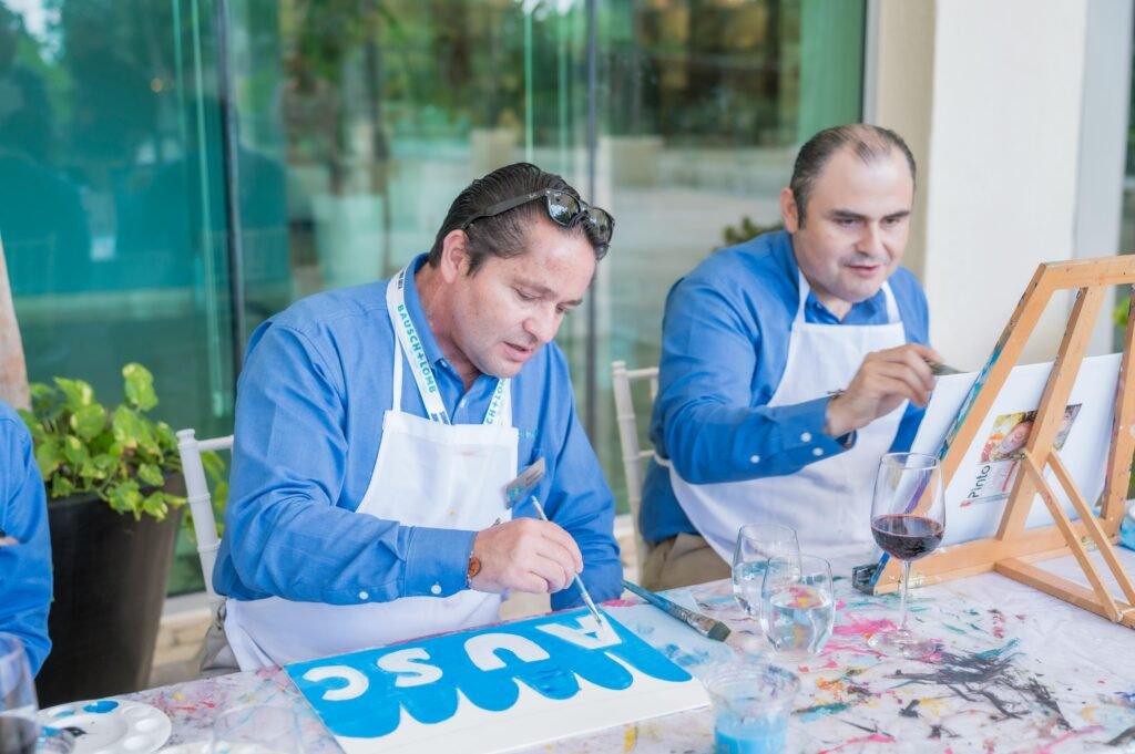 Two men participate in an art workshop, painting on canvases while seated indoors.