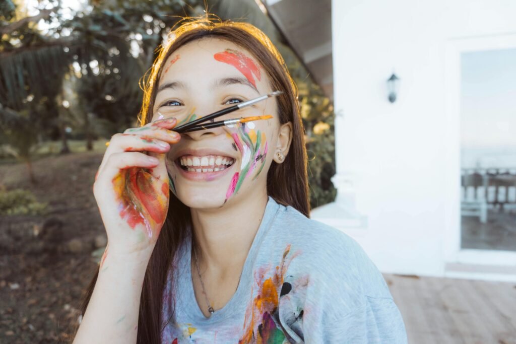 Smiling girl with paint all over her face and brushes held near her nose, enjoying outdoor creativity.