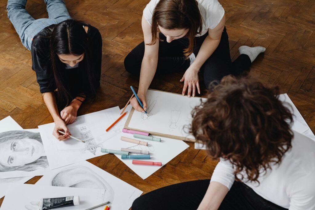 Three women engage in a collaborative drawing session on a wooden floor, surrounded by sketches and art supplies.