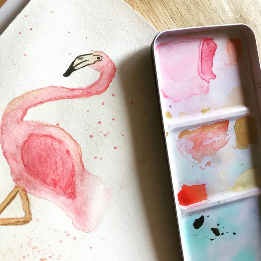 Artistic watercolor flamingo painting beside a colorful paint palette, showcasing creativity and vibrant colors.