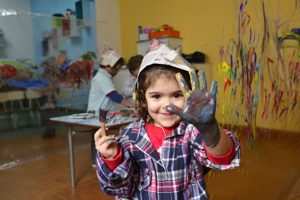 A joyful child paints with colorful paints, wearing a paper hat in an art classroom.